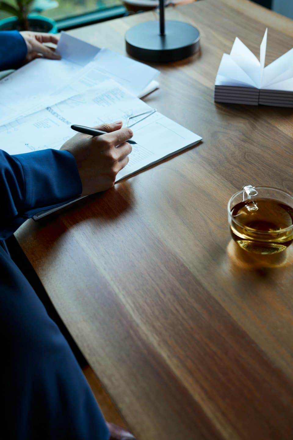 Hands in a blue suit writing on a document at a wooden desk.